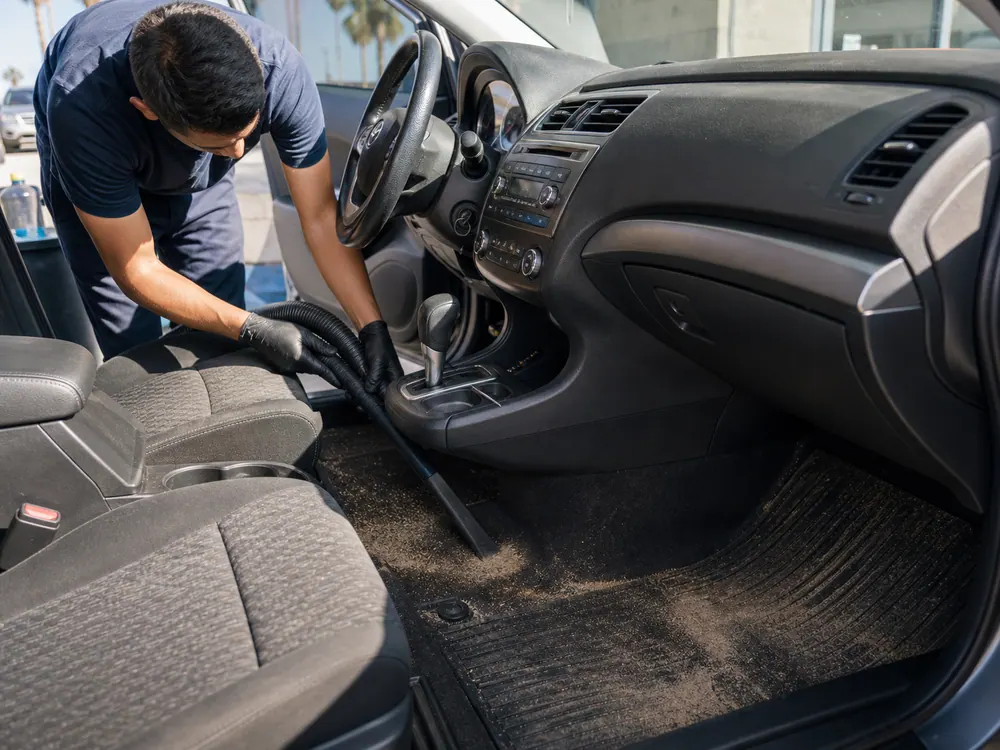Worker vacuuming a sandy car interior and floor mat.